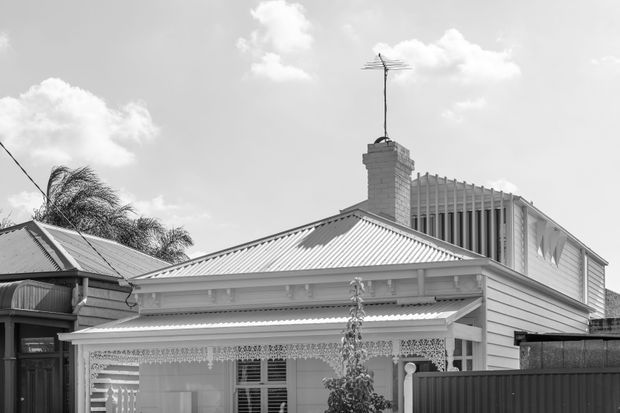 Black-and-white exterior photograph of a traditional single-storey weatherboard house with a corrugated metal gable roof, brick chimney, decorative lacework verandah, timber-framed windows, and a contemporary-looking upper-level extension visible behind the main roofline. The image is composed primarily around the front facade and roof forms.