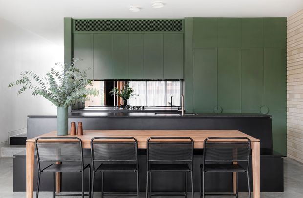 A refined contemporary kitchen and dining area with a long timber dining table and four black dining chairs in the foreground. The kitchen features extensive dark green built-in cabinetry, a dark island bench, stainless-steel sink and tapware, and a black-framed window with sheer curtains. A white painted ceiling and light brick feature wall contrast with the deep green joinery, while a large leafy arrangement adds an organic accent.
