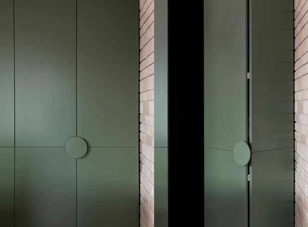 Close-up architectural interior detail showing muted sage-green flush wall or door panels with precise vertical and horizontal joints and circular integrated pulls. Pale exposed brick piers frame a deep black recessed opening, while a narrow reflective glass or dark-framed panel appears on the right. The composition is minimalist, geometric, and softly lit.