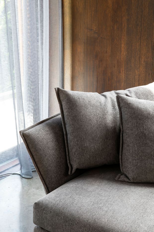 Close-up interior detail of a contemporary upholstered sofa with several grey-brown cushions, positioned beside sheer floor-to-ceiling curtains and a dark timber wall panel. A polished concrete floor is visible at the lower left, with soft natural light creating a calm, understated atmosphere.