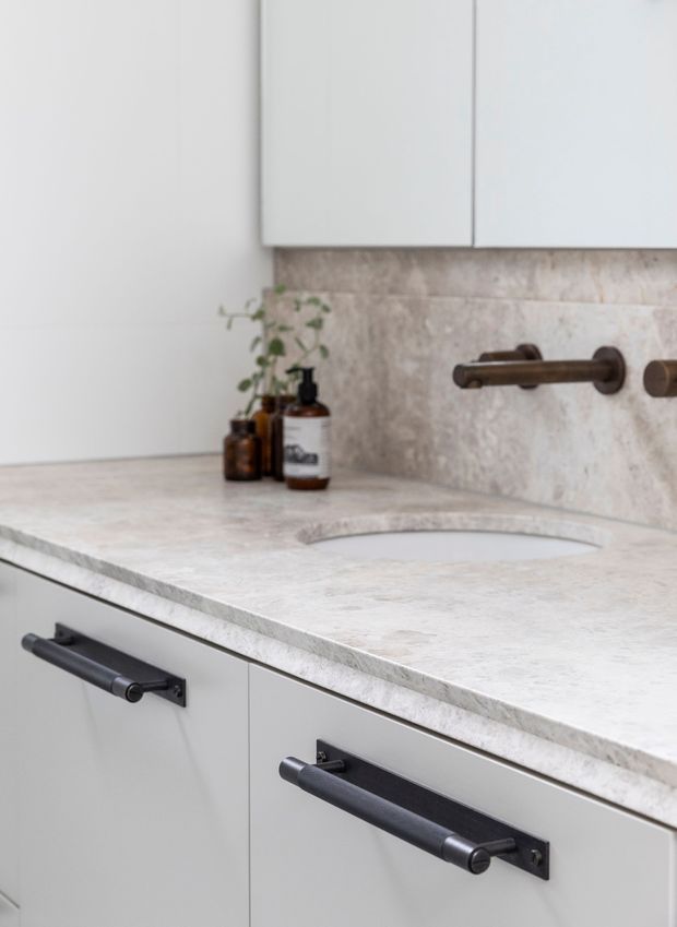 Close-up of a refined contemporary bathroom vanity with white flat-panel cabinetry, a pale natural-stone countertop and matching backsplash, an integrated curved basin, and dark bronze or black hardware and wall-mounted tapware. Small amber toiletry bottles and a trailing plant sit on the counter.