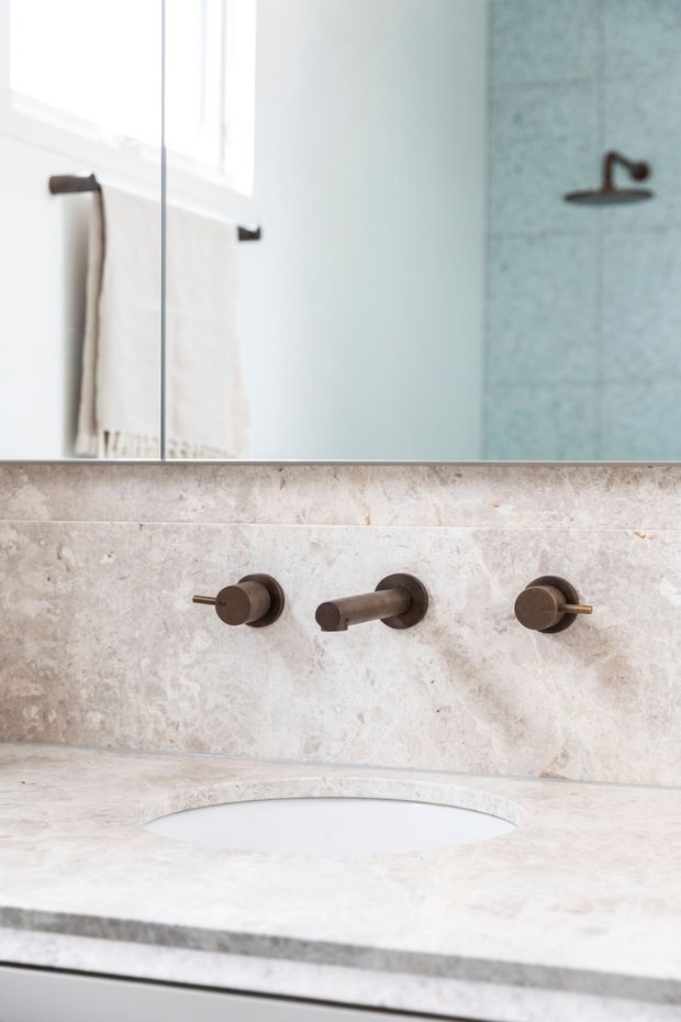 Close-up of a refined bathroom vanity with a pale natural stone benchtop and matching stone backsplash, integrated oval basin, wall-mounted bronze tapware, and a large mirror reflecting a towel rail and shower wall. The space is bright, minimal, and softly neutral with warm metallic accents.