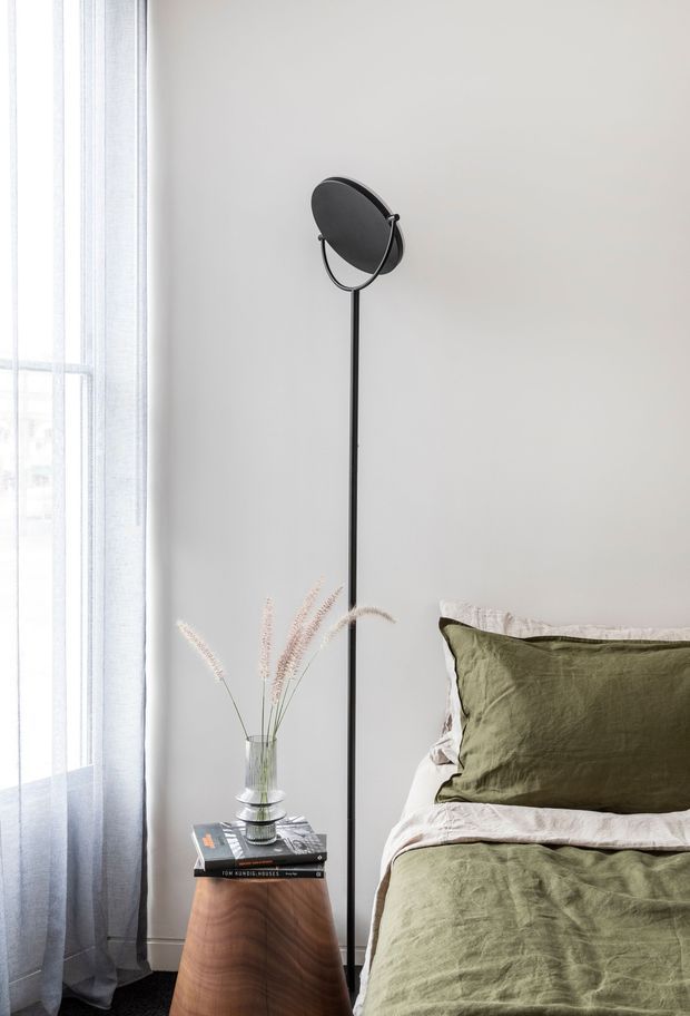 A softly lit bedroom corner with a low bed dressed in muted olive-green linen and off-white bedding, beside a compact curved timber bedside table. A tall slender black floor lamp with a circular shade stands against a light neutral wall, while sheer curtains filter daylight from a large window. Minimal styling includes stacked books and a glass vase with dried grasses.