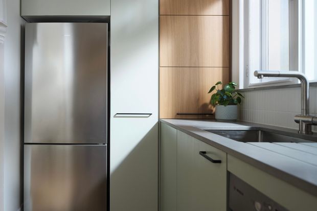 Compact contemporary kitchen with a stainless-steel refrigerator, pale green integrated cabinetry, warm timber wall cabinetry, a grey tiled splashback, and a dark benchtop surrounding a stainless-steel sink. Natural light enters through the window above the sink, with a small potted plant adding greenery.