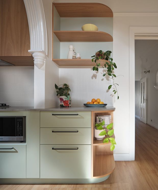 A refined kitchen interior with pale sage-green cabinetry, black bar handles, a white vertically textured tile splashback, and warm timber flooring. Open curved timber shelving displays ceramics and plants, while a rounded timber countertop end and decorative fluted architectural trim add character. A hallway is visible beyond the kitchen.