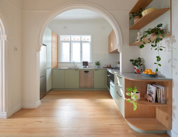 A bright, compact kitchen viewed through a prominent curved plaster archway. The room features pale green flat-panel cabinetry, a stainless-steel dishwasher, light stone benchtops, timber flooring, open timber shelving with plants and books, and a small window above the rear work area. The arched opening and decorative wall detailing add a refined traditional character, while the cabinetry and joinery are contemporary.