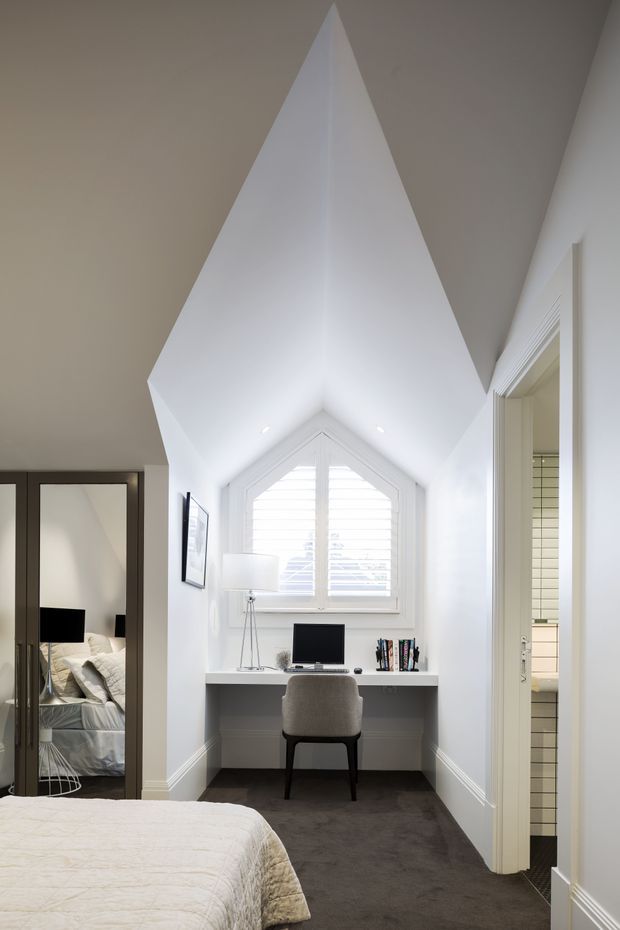 A compact, bright bedroom nook with a built-in white desk positioned beneath a small gabled window with plantation shutters. The room has a dramatic white vaulted and angular ceiling, grey carpet, white walls and trim, a simple upholstered desk chair, and a mirrored wardrobe door reflecting the bed. A bathroom doorway is visible on the right.