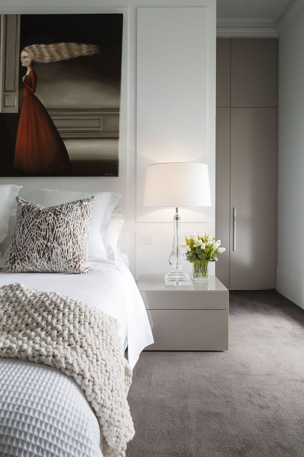 A refined bedroom interior featuring a neatly made bed with white textured bedding, a chunky cream throw, and a patterned accent cushion. A minimalist grey bedside table holds a clear glass lamp and flowers, while a tall built-in grey wardrobe provides storage. The room has pale walls with decorative moulding, plush grey carpet, and a large framed artwork above the bed.