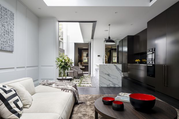 A bright, open-plan contemporary living and kitchen area. The foreground features a light upholstered sofa with patterned cushions and a dark oval coffee table, while the rear contains a compact marble-look kitchen island, dark full-height cabinetry, integrated appliances, and a dining or sitting area beyond. White wall panelling, a large skylight or roof opening, pendant lighting, and a patterned rug complete the refined interior.