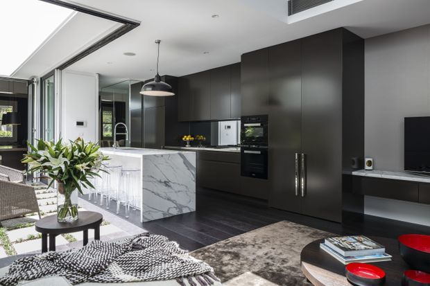 Open-plan contemporary kitchen and living area with dark matte cabinetry, a veined white stone island, integrated appliances, pendant lighting, and dark flooring. The kitchen opens to a bright glazed outdoor area, while a lounge zone with a television, coffee table, rug, armchair, and soft furnishings occupies the foreground.