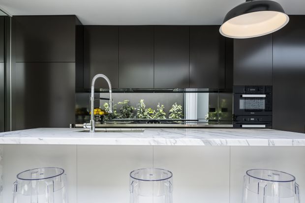 Contemporary kitchen featuring a large white marble-look island with integrated seating, dark matte cabinetry, reflective splashback, stainless steel gooseneck faucet, built-in black appliances, and a prominent round pendant light. Three transparent bar stools line the island.