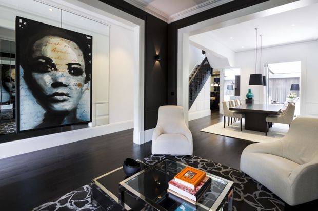 Contemporary open-plan living and dining area with dark timber flooring, black-and-white feature walls, large monochrome artwork, sculptural lounge chairs, a glass coffee table, and a formal dining setting beyond. A staircase and bright white joinery contribute to the refined, high-contrast interior.