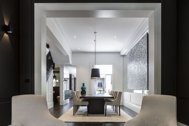 A refined contemporary dining room viewed through a wide interior opening, with a dark-framed portal contrasting against white architectural mouldings and walls. The room features a central dark dining table, upholstered dining chairs, a large black pendant light, dark timber flooring, a pale rug, and a large monochrome textured wall artwork. White-framed glazing and a glimpse of a stair or adjoining living area are visible beyond.