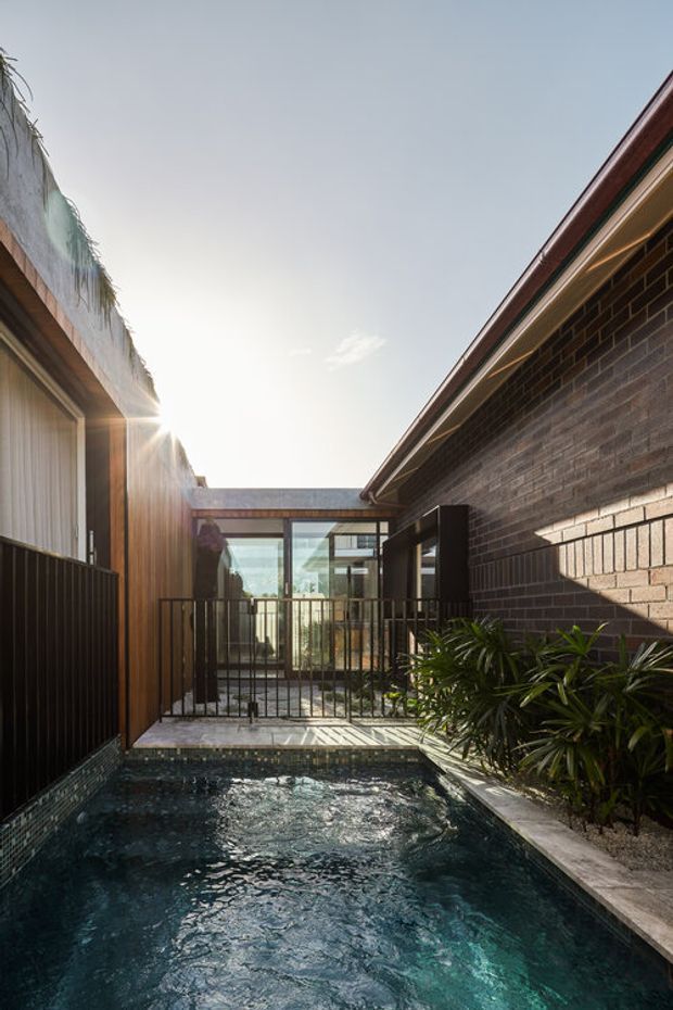 A narrow courtyard pool framed by contemporary residential architecture. Dark brick walls, warm vertical timber cladding, black metal railings, and glazed doors surround the reflective blue-green water. Strong sunlight enters from above, creating a calm, private outdoor space with dense tropical planting along one edge.