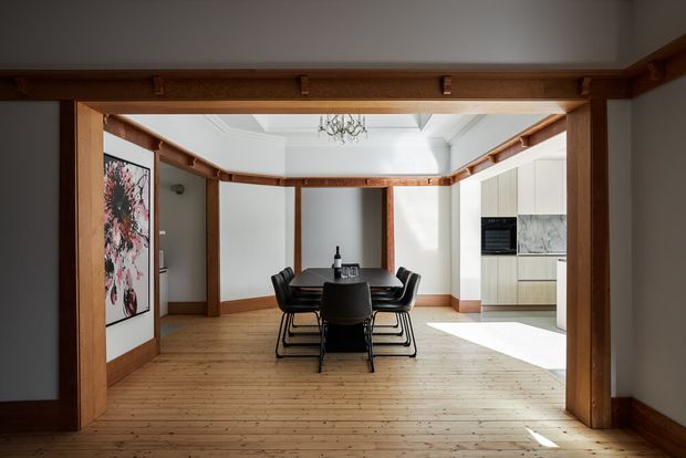 A bright open-plan dining area viewed through a substantial timber-framed opening. The room features a rectangular dark dining table with black chairs, wide timber floorboards, white walls and ceiling, exposed timber beams and trims, and a contemporary white kitchen with pale stone-look surfaces in the background. A decorative chandelier and framed artwork add restrained detail.