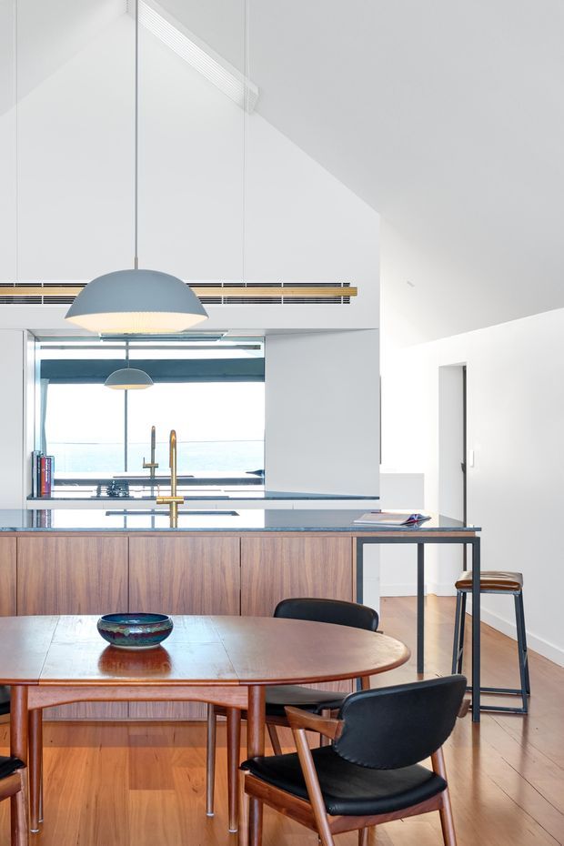 Bright open-plan kitchen and dining area with a high angled white ceiling, timber kitchen island cabinetry, stone benchtops, timber flooring, and a mid-century-style oval dining table with leather chairs. A large pendant light hangs above the dining area, while a reflective splashback or glazing opens the space toward a bright exterior view.