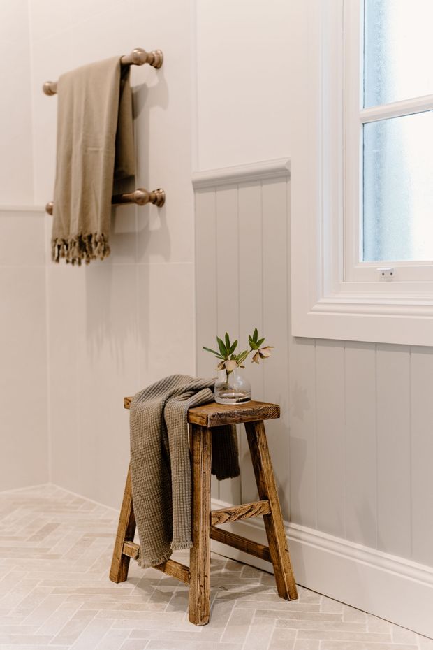 A softly lit, refined bathroom corner featuring pale tiled walls, vertical painted wall panelling, white trim, and a frosted window. A brass towel rail holds a neutral fringed towel, while a rustic timber stool displays another folded throw and a small glass vase with greenery. The floor is finished in a light herringbone tile pattern.