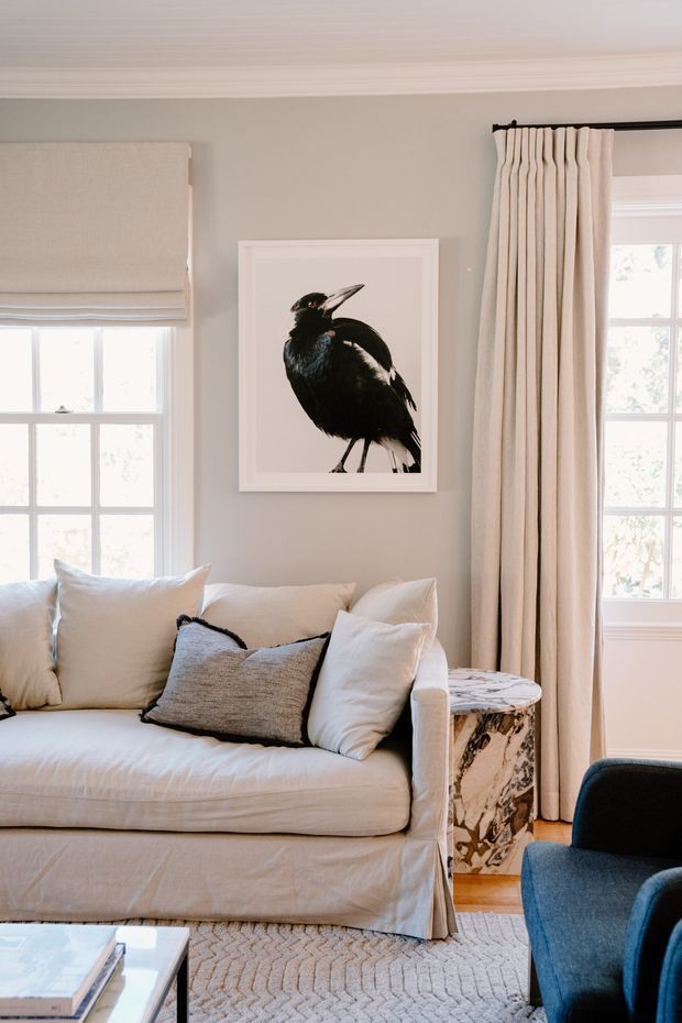 Bright, softly styled living room with a white slipcovered sofa layered with neutral cushions, a textured grey accent pillow, pale grey walls, white trim, and large divided-light windows dressed with Roman and full-length curtains. A framed black bird artwork hangs above a marble-patterned side table, with a blue armchair and pale textured rug completing the space.