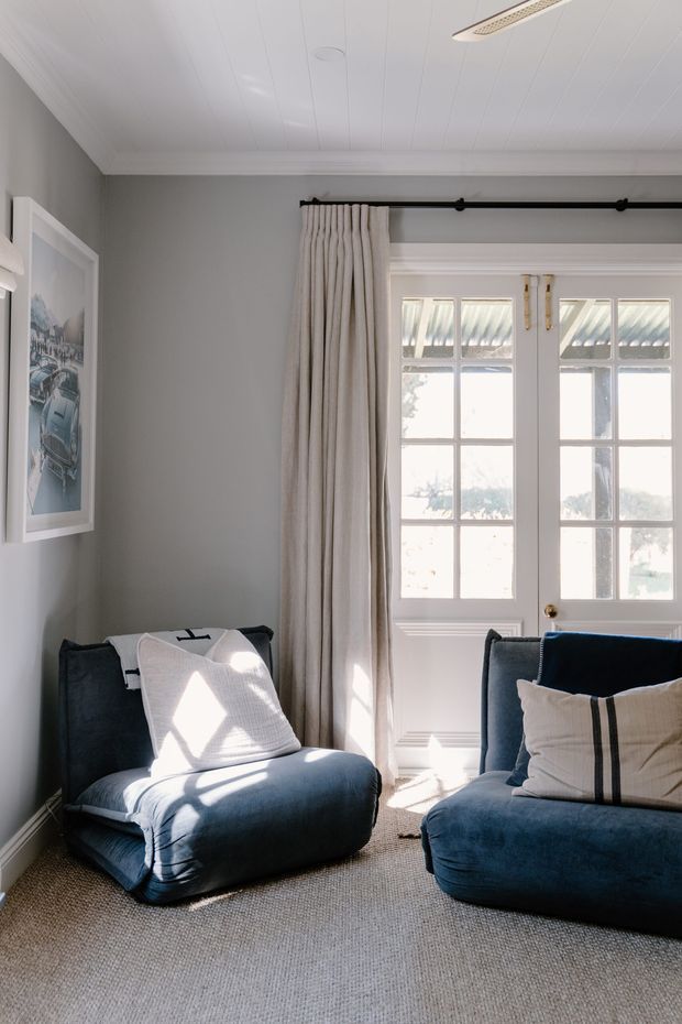 A bright, cozy sitting area with deep blue upholstered lounge chairs and sofa, neutral cushions, full-height cream curtains, and white French doors with multiple glass panes. The room has grey painted walls, a white tongue-and-groove style ceiling, beige textured carpet, and framed artwork. Sunlight creates strong geometric patches across the seating and floor.
