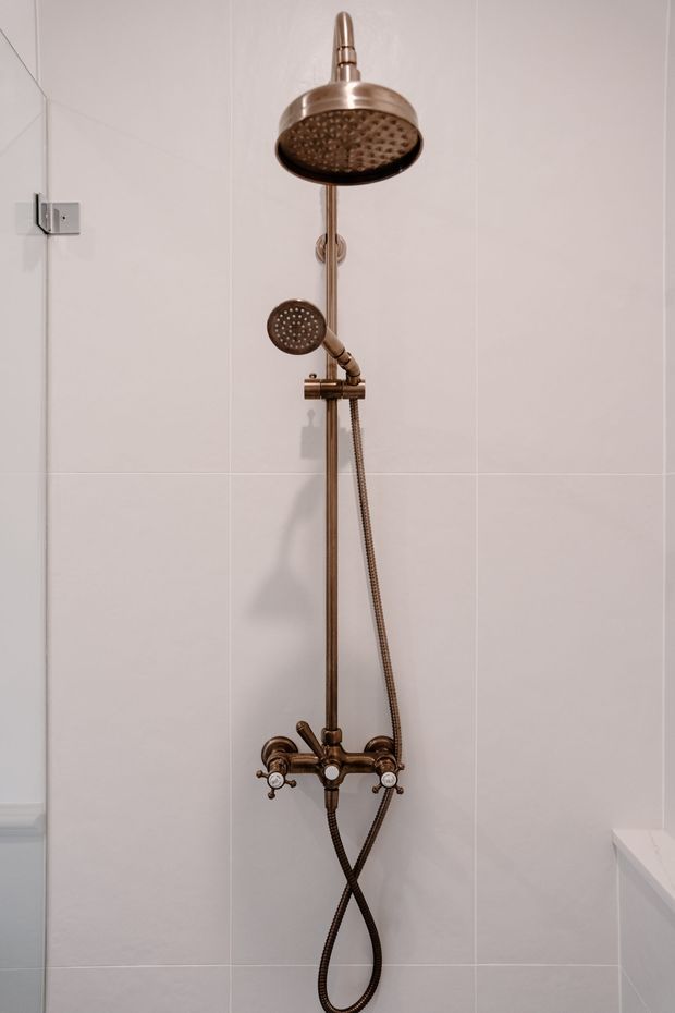 Close-up view of a bathroom shower wall featuring a vintage-style brushed bronze or copper-toned exposed shower system. The installation includes a large round overhead rain shower, handheld shower on a vertical rail, flexible hose, and traditional thermostatic controls. The shower is mounted against large pale grey-white tiled walls, with a partial glass shower panel visible at left and a white built-in ledge or fixture at right.