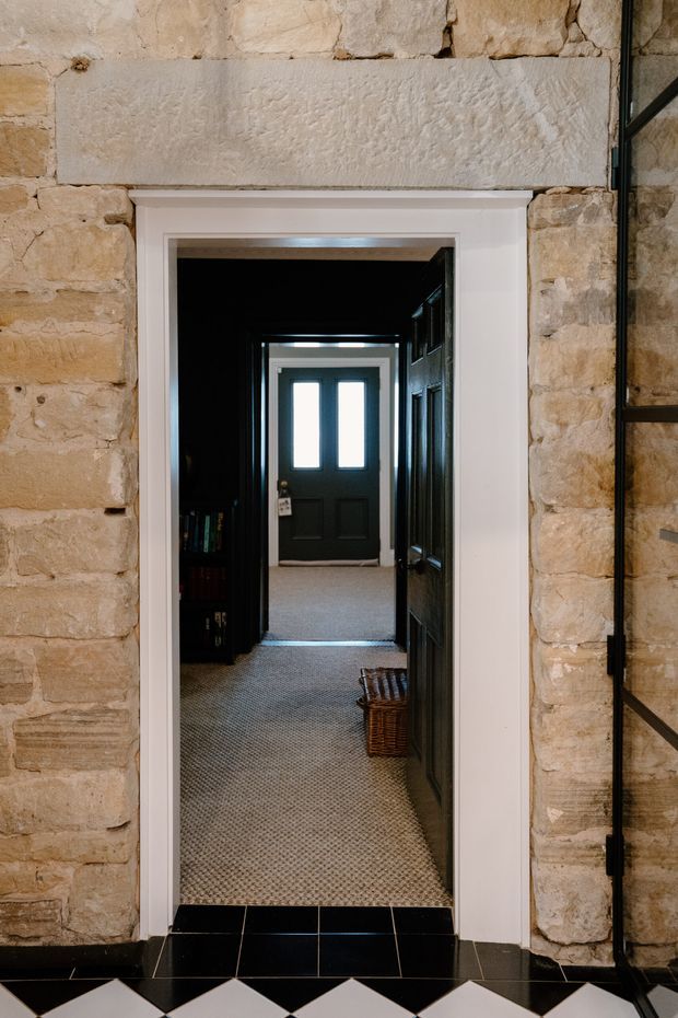 A narrow interior doorway connects a dark tiled foreground space to a carpeted hallway. The opening has crisp white trim set into a rustic exposed stone wall, with a dark paneled door and glazed exterior-style door visible deeper in the corridor. Black-and-white geometric floor tiles appear in the foreground, while a woven basket and shelving add modest furnishing detail.