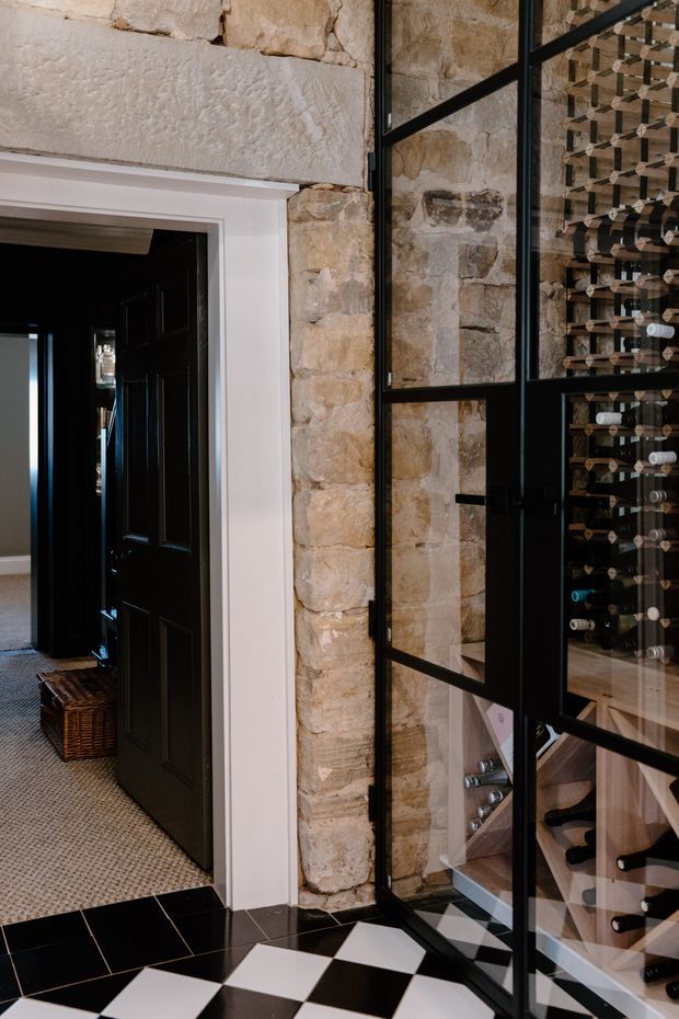 Interior view of a refined wine cellar or wine storage area behind black metal-framed glass doors, set into an exposed natural stone wall. The foreground has a bold black-and-white checkerboard tile floor, while the adjacent hallway features dark timber doors, white trim, and a textured neutral carpet. Wine bottles are displayed in extensive built-in timber racks.