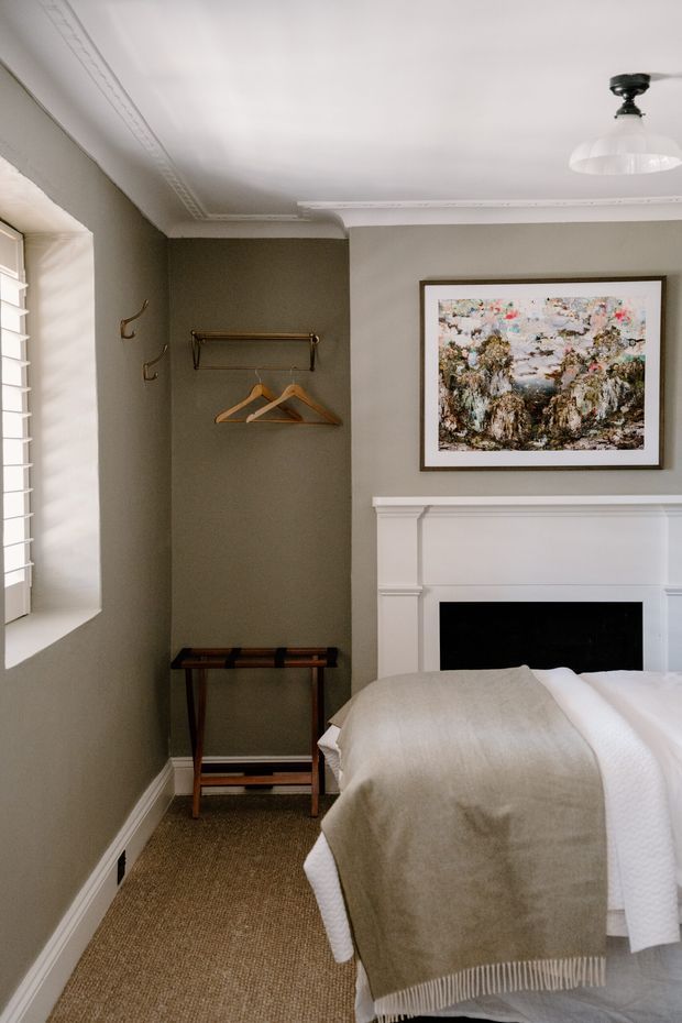 A compact, traditionally detailed bedroom with muted taupe walls, a white decorative cornice and fireplace mantel, and a partially visible neatly made bed. A framed abstract artwork hangs above the fireplace, while a simple timber-and-metal clothes rail, hooks, and console occupy the alcove beside a louvred window. The room has warm textured carpet, classic trim, and a vintage-inspired ceiling pendant.