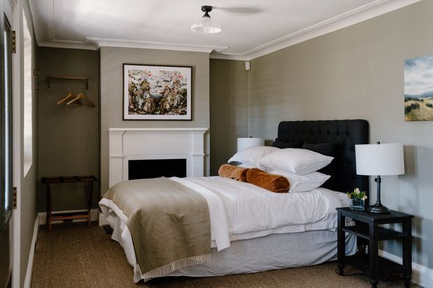 A refined bedroom with a large upholstered bed, white layered bedding, a taupe throw, black bedside table and lamp, and a dark tufted headboard. The room has muted grey-green walls, traditional white cornices and mouldings, a classic white fireplace mantel, framed artwork, and a small open hanging area near the doorway. Carpeted floor and soft natural light create a calm, elegant atmosphere.