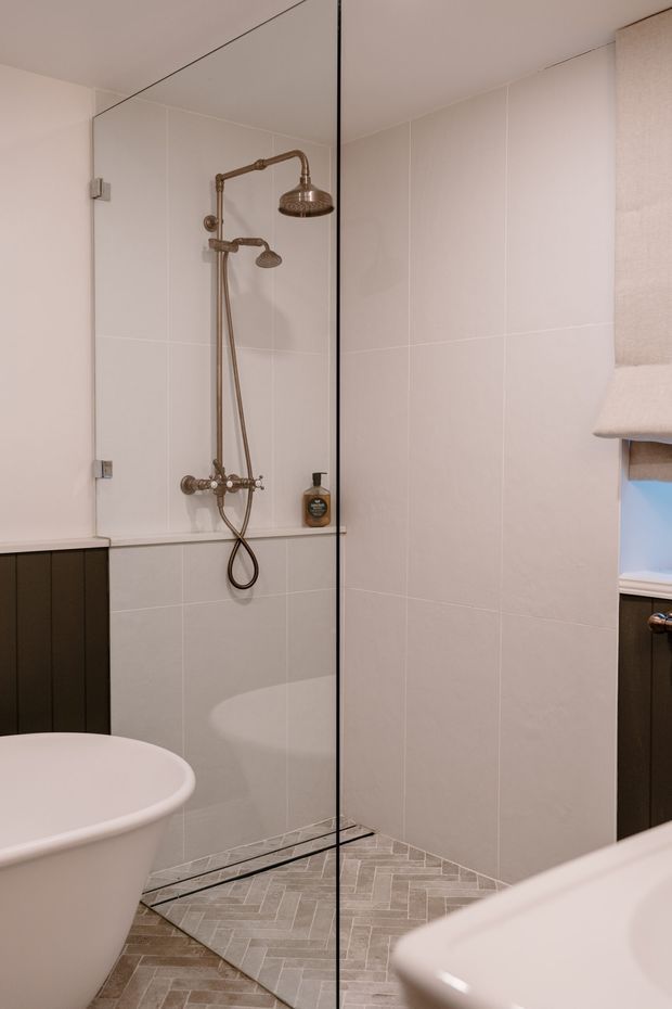 A compact, refined bathroom featuring a frameless glass walk-in shower with dual antique-bronze shower fittings, pale large-format wall tiles, and a light grey herringbone tiled floor. A freestanding white bathtub is partially visible in the foreground, with dark vertical-panel cabinetry and a small window with a fabric blind at the side.