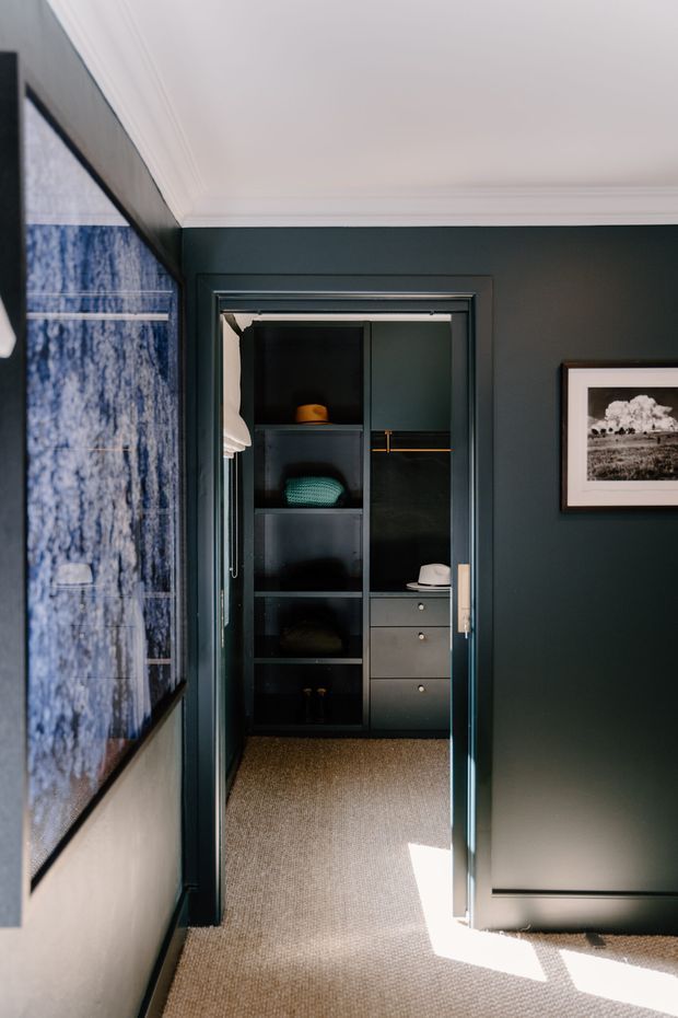 A refined dark-toned dressing room or walk-in wardrobe viewed through an open doorway. The space features deep green built-in cabinetry with open shelving, drawers, hanging storage, and brass detailing, set against a dark green wall with white decorative ceiling cornices. Beige carpet, framed artwork, and a large blue-toned artwork or textile contribute to the tailored, moody interior.