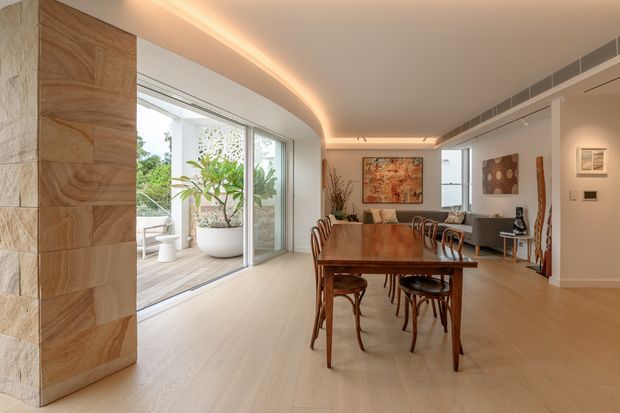 Open-plan contemporary dining and living area with a long timber dining table and curved-back chairs in the foreground, flowing into a lounge at the rear. Full-height glazing opens to a planted outdoor terrace, while a curved recessed ceiling with concealed warm lighting runs through the space. The interior features pale timber flooring, warm timber wall cladding, white walls, artwork, and restrained modern furnishings.