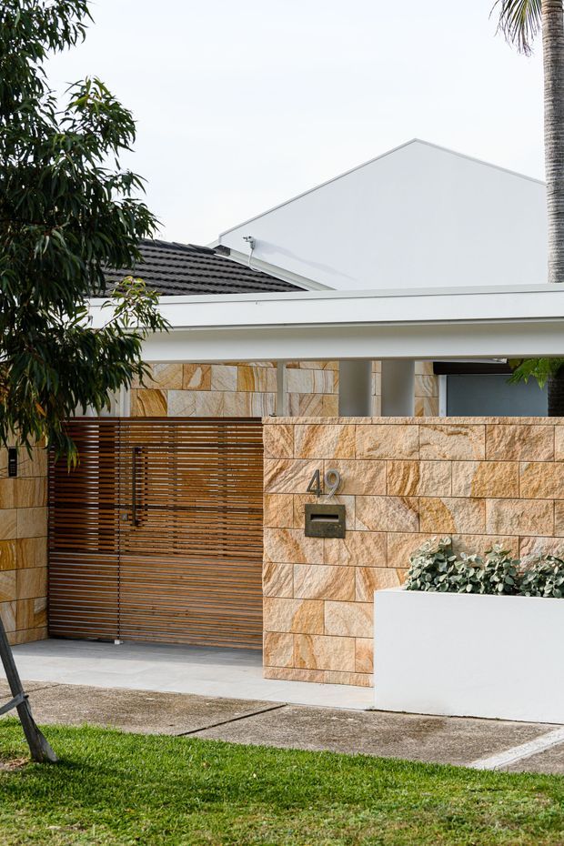 Street-facing view of a contemporary residential entry and carport. The frontage features a warm sandstone block wall with house number and letterbox, a horizontal timber slatted gate, white rendered walls, a broad flat-roofed canopy supported by simple round columns, and a tiled pitched roof behind. Landscaping includes a raised white planter and mature greenery.