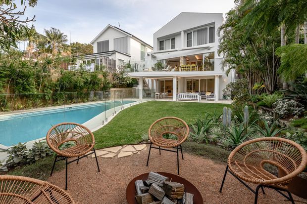 A landscaped residential backyard with a contemporary multi-level white house, expansive covered terraces, a swimming pool, and a gravel outdoor seating area with woven lounge chairs surrounding a fire bowl. Dense tropical planting frames the garden and pool, while a glazed pool barrier and open lawn connect the outdoor spaces.