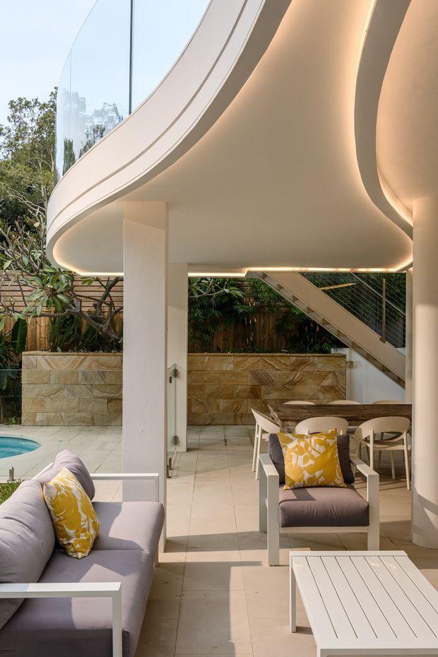 A refined outdoor living and dining area beneath a sweeping curved architectural canopy, with integrated warm lighting, white structural columns, and a tiled terrace. The space includes white-framed lounge furniture with pale upholstered cushions and yellow patterned pillows, a dining table and chairs, a swimming pool edge, landscaped planting, a stone privacy wall, and a stairway with metal balustrade. The setting has a light, resort-like contemporary feel.