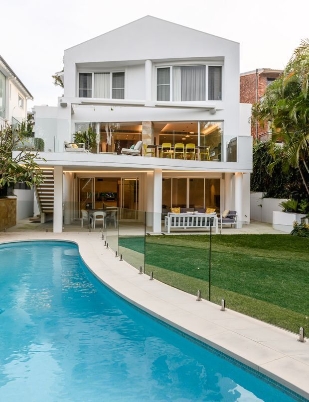 Rear exterior view of a contemporary multi-storey white house with a simple gable roof, extensive glazing, balconies and open-plan entertaining areas overlooking a swimming pool. A glass pool fence, landscaped lawn, palms and outdoor seating occupy the foreground and garden.