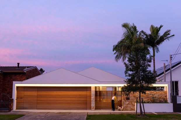 Single-storey contemporary residential facade photographed at dusk, featuring a broad timber-clad garage door, warm illuminated entry and glazing, pale rendered walls, and a stone-clad entry section. Palm trees and neighbouring homes frame the suburban streetscape.