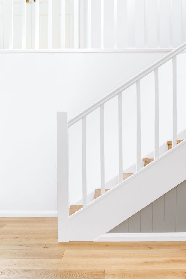 Bright, minimalist interior view of a white-painted staircase with simple vertical balusters, white handrail and stair stringer, and light timber treads. The staircase rises diagonally across a white wall with subtle vertical panel detailing, while pale timber flooring continues through the lower level.