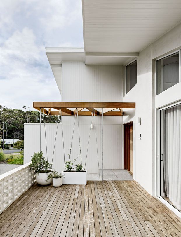 Bright exterior view of a contemporary two-storey residence focused on a spacious timber deck and covered entry terrace. The facade combines white rendered and vertically panelled walls, broad white-framed glazing, a flat projecting roof, and a timber pergola-like canopy with suspended cables. Potted plants and a breeze-block balustrade add detail, with a leafy suburban setting beyond.