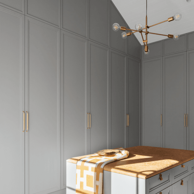 A refined walk-in wardrobe or dressing room with full-height grey paneled cabinetry, slim brass handles, and a central island with a warm wood benchtop and drawers. A sculptural brass multi-arm pendant with globe bulbs hangs overhead, casting strong geometric shadows across the island. The space has a tailored, luxurious and modern-classic appearance.