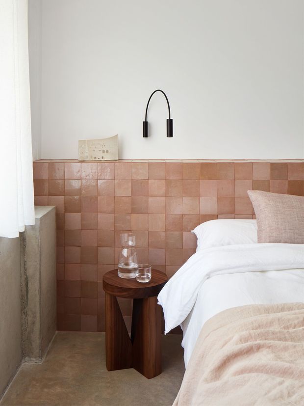 A softly styled bedroom corner with a bed dressed in white linen and a pale beige throw, positioned beside a dark timber bedside table. The room features a half-height wall of glossy terracotta-pink square tiles, warm concrete flooring, white plaster above, a black arched wall light, and a sheer white curtain. The overall composition is warm, minimal, and tactile.