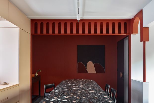A compact, richly coloured dining room with a long rectangular dining table in a dark patterned stone finish, surrounded by black chairs. The room features deep red painted walls and a striking red built-in architectural screen with repeated rounded vertical openings above the rear wall. A dark abstract artwork, concealed cabinetry, a tall doorway, and a linear ceiling light complete the contemporary interior.