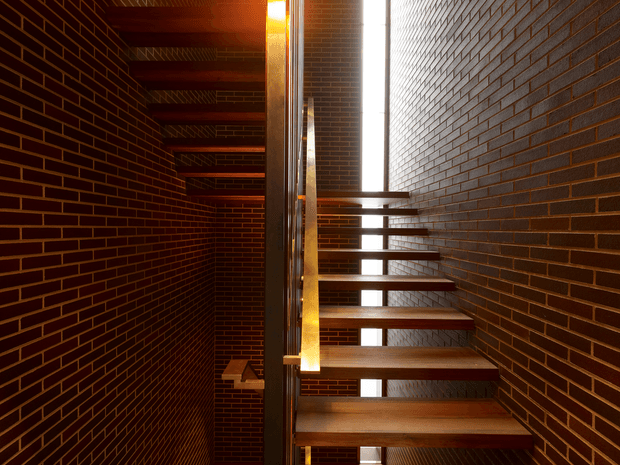 Interior view of a narrow, dramatic stairway enclosed by dark exposed brick walls. Open-riser timber stairs are supported by a central dark metal structure, with tall vertical glazing bringing in strong daylight along the stairwell. The brickwork continues across the ceiling and surrounding walls, creating a warm, textural, minimalist-industrial atmosphere.