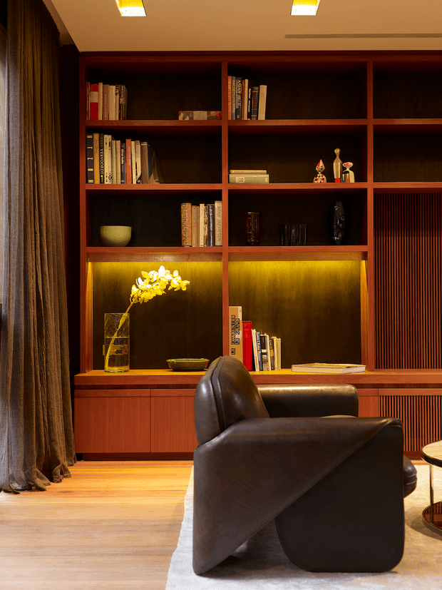 A warmly lit living room featuring a full-height dark timber built-in shelving and cabinetry wall, with books, decorative objects, integrated golden accent lighting, and vertical slatted panels. A sculptural dark brown leather lounge chair sits in the foreground on a light timber floor, beside floor-length curtains and a partially visible round side table. The space has a rich, refined, mid-century-inspired interior with strong custom joinery.