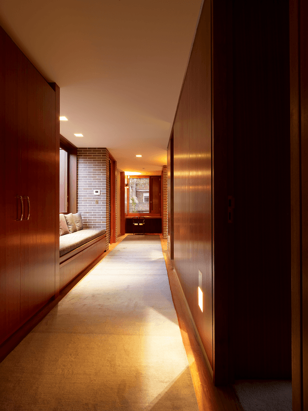 A long, warm-toned residential hallway with dark timber wall panelling and built-in cabinetry, a pale carpeted floor, recessed ceiling lights, and a brick-lined window seat on the left. The corridor leads toward a small timber-framed window and built-in joinery at the far end, creating a narrow, intimate and strongly linear composition.