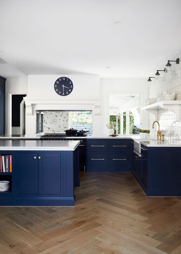 Bright, refined kitchen with deep navy shaker-style cabinetry, pale stone benchtops, glossy white tiled splashback, brass tapware and hardware, and a warm timber herringbone floor. The room includes a large island, generous fixed storage, open shelving, a classic mantel-like rangehood surround, and abundant daylight.