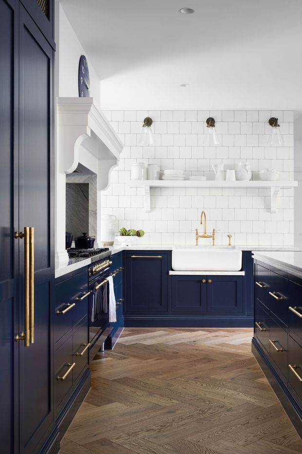 A refined galley kitchen with deep navy-blue shaker cabinetry, brass handles and fixtures, a white farmhouse-style apron sink, and a white subway-tile backsplash. Open shelving displays white ceramics, while a classic range sits beneath a decorative white mantel-style hood surround. The floor is laid in a warm natural timber herringbone pattern, creating a traditional yet polished interior.