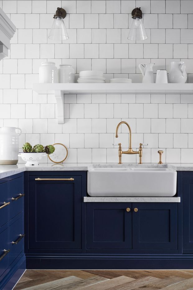 A refined, traditional-style kitchen featuring deep navy-blue shaker cabinetry with brass hardware, a white farmhouse apron-front sink, marble benchtops, and a white rectangular subway-tile splashback. Open white shelving displays ceramics, while brass wall sconces and a gooseneck tap add warm metallic accents. The floor is laid in a geometric chevron or herringbone timber pattern.