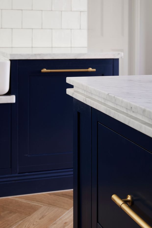 Close-up view of a refined kitchen featuring deep navy-blue shaker cabinetry, white marble countertops with layered edge detailing, brass bar and drawer pulls, white rectangular backsplash tile, and warm herringbone timber flooring.