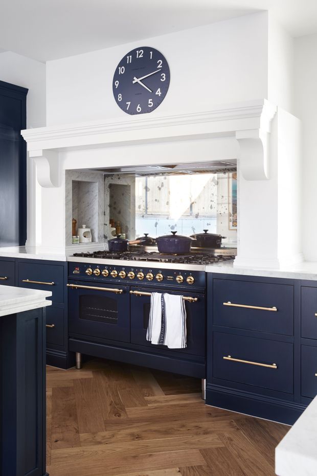 A bright, traditional-style kitchen centred on a large navy range cooker with brass controls and handles. Deep navy cabinetry with brass pulls surrounds the cooker, while white stone benchtops, white walls, and a decorative white mantel-style rangehood surround create a strong contrast. A mirrored splashback reflects the room, and warm timber herringbone flooring adds texture. A dark wall clock is mounted above the mantel.