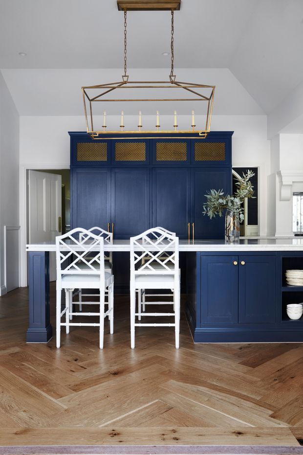 A refined kitchen interior with deep navy-blue floor-to-ceiling cabinetry, brass mesh upper cabinet inserts and hardware, white marble-look benchtops, and two white lattice-back counter stools. A large geometric brass pendant hangs above the island, while a herringbone timber floor and white architectural trim add warmth and classic detail.